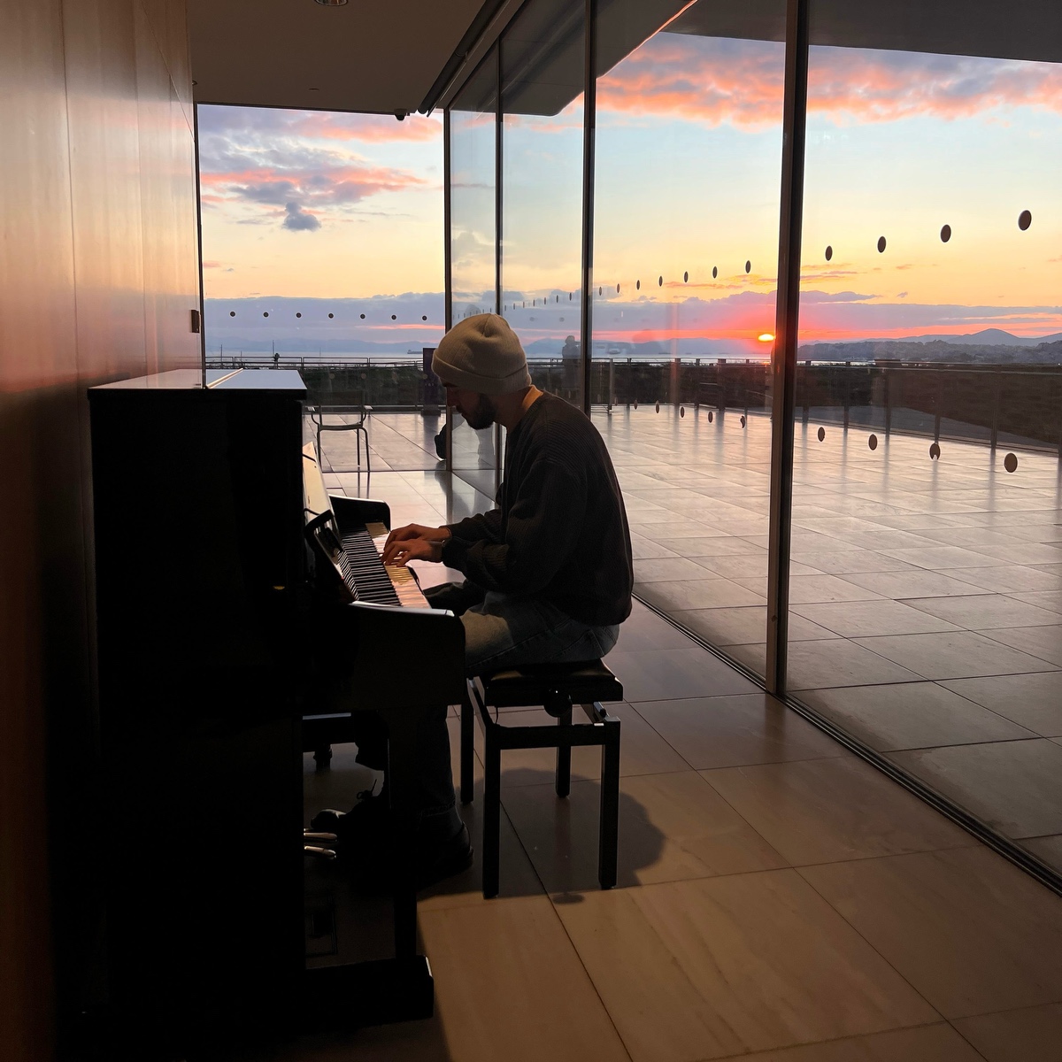 Christos Anagnostou Daly at an upright piano at sunset, beside a glass wall overlooking the sea